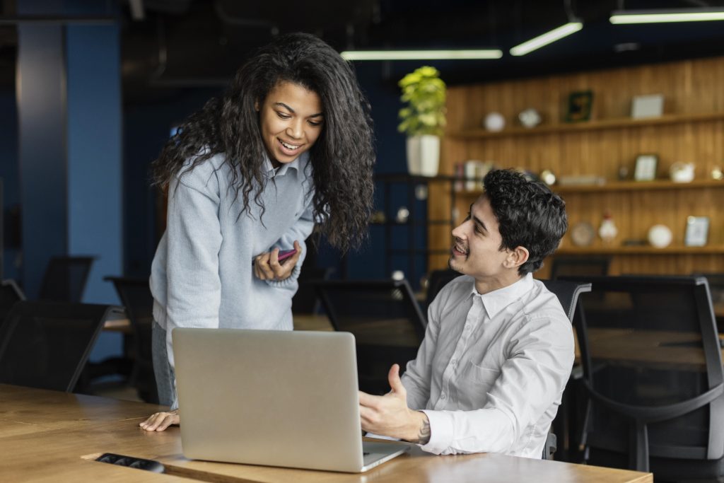 A imagem tem duas pessoas, uma delas esta sentado em frente a um computador olhando para a tela, mostrando para a segunda pessoa que esta em pé. Representando o que é programas de treinamento