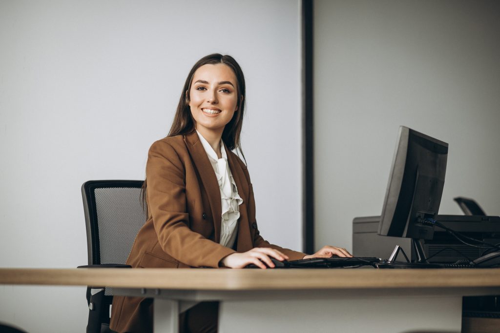 na imagem tem uma mulher sorrindo em frente a um computador em um escritório com caderno sobre a mesa. Representando o que é desenvolvimento profissional